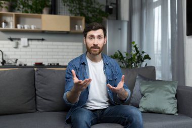 Konsantre olmuş bir adam kameraya bakıyor, görüntülü konuşuyor, evdeki kanepede oturuyor.