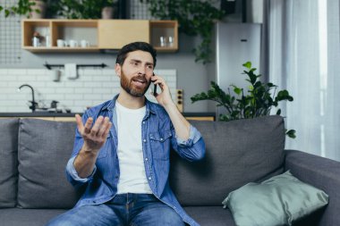 Evdeki adam kanepede oturup telefonla konuşuyor, neşeli ve odaklanmış.