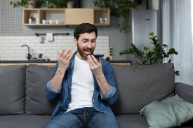 Kızgın adam sesli mesaj kaydediyor, uygulamayı kullanıyor, ve telefonda diktafon kullanıyor, evdeki kanepede oturuyor.