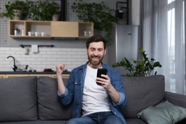 Evdeki bir adam kazandı ve internetten haberlerle yüzleşiyor, telefon ekranına bakıyor ve zaferin zaferini telefonla kutluyor.