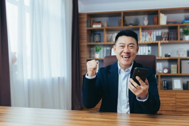 Başarılı bir Asyalı, klasik bir ofiste çalışıyor, elinde telefonla kameraya bakıyor, zaferi kutluyor, başarıyı işaret ediyor.