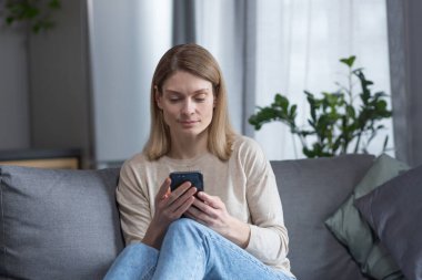 Evdeki kanepede oturan orta yaşlı güzel bir kadın, telefonu kullanıyor ve gülümsüyor.
