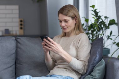 Evdeki kanepede oturan orta yaşlı güzel bir kadın, telefonu kullanıyor ve gülümsüyor.