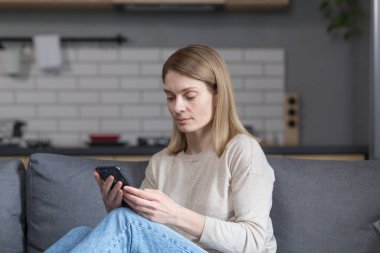 Evdeki kanepede oturan orta yaşlı güzel bir kadın, telefonu kullanıyor ve gülümsüyor.