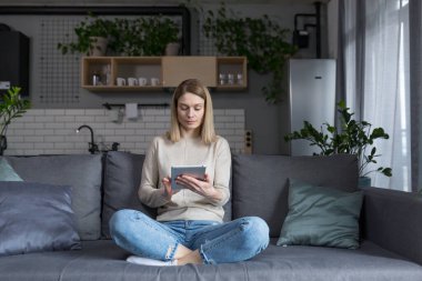 Evdeki kanepede oturan, tablet bilgisayar kullanan, e-kitap okuyan ve internetten öğrenen bir kadın.