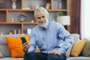 Mutlu yaşlı adam evdeki telefonu kullanıyor koltukta oturuyor, mobil uygulamalar kullanıyor.