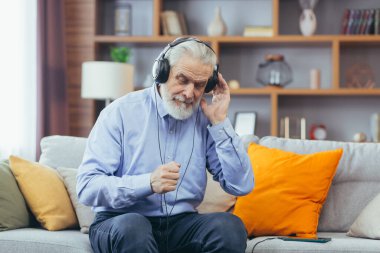 Evde koltukta oturan, kulaklıkla müzik dinleyen, telefonda müzik uygulaması kullanan yaşlı, kır saçlı adam.