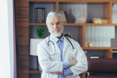 Deneyimli bir doktorun portresi, kliniğin baş gri saçlı doktoru, ofisinde, çapraz kollarla kameraya bakıyor.