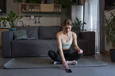 Evde yoga yapan bir kadın lotus pozisyonunda yerde oturuyor, telefonu online fitness ve yoga dersleri için kullanıyor.