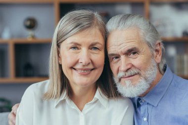 Yakın plan mutlu, kır saçlı evli bir çiftin portresi. Evde dikilirken kameraya bakıyorlar. Kapalı alanda. Karı koca gülümseyen emekliler. Yaşlı insanlar sarılır. Neşeli büyükanne ve büyükbaba kaynaşıyor