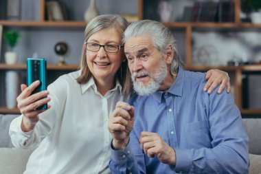 Yaşlı gri saçlı evli çift, evde birlikte video oyunu oynuyorlar, koltukta oturuyorlar, telefon kullanıyorlar ve internet üzerinden görüşüyorlar.
