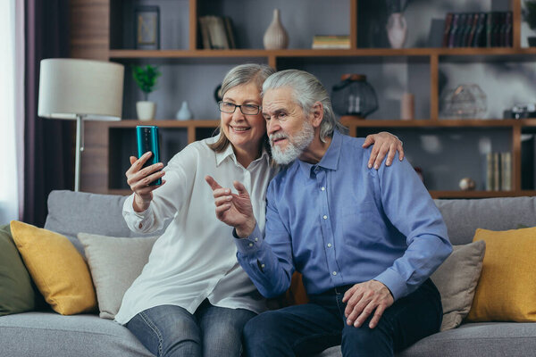Senior gray-haired married couple man and woman at home together, play video call, sit on sofa at home, use phone for call and online meeting