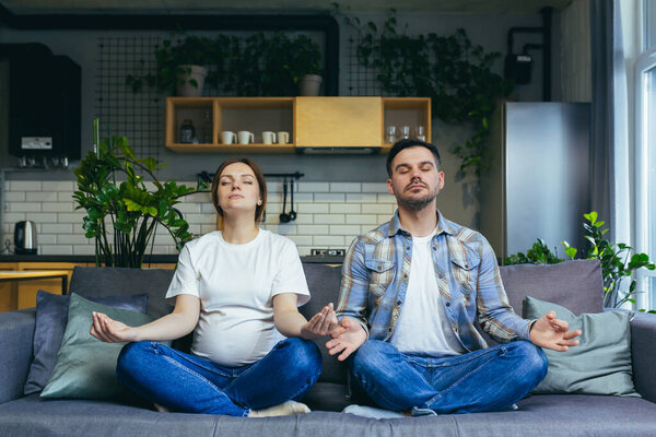 A pregnant man and woman are meditating in a lotus position on the couch