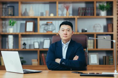 Ciddi bir Asyalı ofiste oturmuş kolları bağlı kameraya bakıyor, işadamı patronunun portresi.