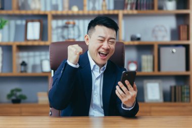 Başarılı bir Asyalı klasik bir ofiste çalışıyor, elinde telefon, zaferi kutluyor, başarı jesti yapıyor.