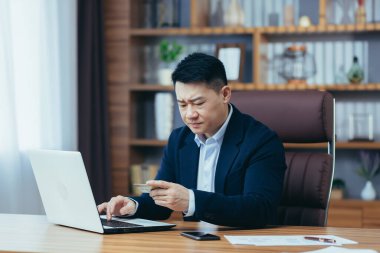 Asyalı üzgün bir adam, online alışveriş yapmaya çalışıyor, ofiste oturuyor, bir kredi kartı kullanıyor, ve bir dizüstü bilgisayar kullanıyor, şifreyi hatırlamaya çalışıyor.