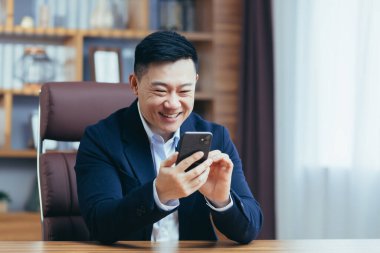 Ofiste çalışan başarılı Asyalı işadamı, masada oturuyor, telefonda yazıyor, mutlu ve gülümsüyor.