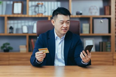 Ofiste otururken mutlu Asya alışverişleri telefon uygulaması ve banka kartı kullanan bir adam online alışverişin tadını çıkaran bir işadamı.