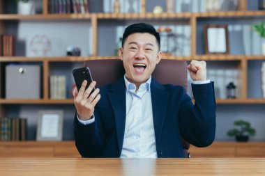 Genç mutlu Asyalı adam bahsi, ödülü, rekabeti kazandı, telefonu kullanıyor. Ofiste, takım elbiseli bir masada oturuyor.