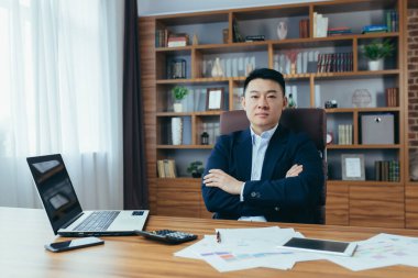 Genç yakışıklı Asyalı bir adamın portresi, iş adamı, takım elbiseli bir patron, ofiste bir masada bir sandalyede oturuyor, bir dizüstü bilgisayar ve evraklarla çalışıyor, kameraya bakıyor. Kollarını çapraz yapmış, ciddi.