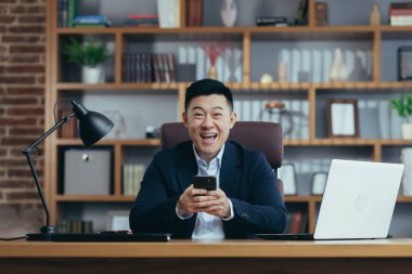 Genç mutlu Asyalı adam bahsi, ödülü, rekabeti kazandı, telefonu kullanıyor. Ofiste, takım elbiseli bir masada oturuyor.