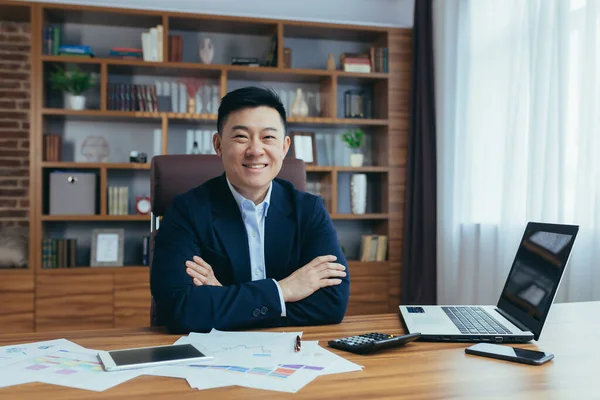 Genç yakışıklı Asyalı bir adamın portresi, iş adamı, takım elbiseli bir patron, ofiste bir masada bir sandalyede oturuyor, bir dizüstü bilgisayar ve evraklarla çalışıyor, kameraya bakıyor. Kollarını kavuşturdu, gülümsedi.