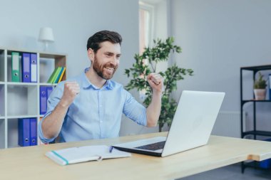 Başarılı serbest çalışan işadamı, merkez büroda çalışır, dizüstü bilgisayara bakar ve zaferi kutlar, el ele tutuşur ve gülümser, zafer jesti yapar.