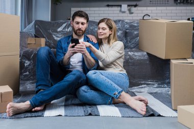 Yeni bir dairede bir adam ve bir kadın, yerde oturan evli bir çift, karton kutuların yanında, telefonu online alışveriş için kullanıyorlar, mobilyaları seçiyorlar.