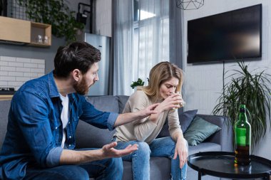 Kadın alkolizmi, evde aile kavgası, içki içen eş, onu durdurmaya çalışan koca, kanepede oturuyor.