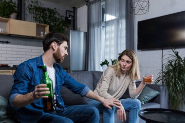Kadın alkolizmi, evde aile kavgası, içki içen eş, onu durdurmaya çalışan koca, kanepede oturuyor.