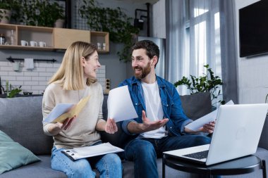 Mutlu evli çift evde evrak işleriyle uğraşıyor, aile bütçesini hesaplıyor, faturaları ödüyor, dizüstü bilgisayar kullanıyor, evdeki kanepede oturuyor.
