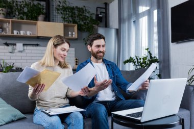 Mutlu evli çift evde evrak işleriyle uğraşıyor, aile bütçesini hesaplıyor, faturaları ödüyor, dizüstü bilgisayar kullanıyor, evdeki kanepede oturuyor.