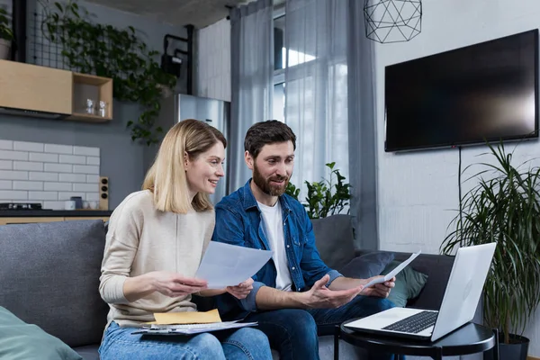 Mutlu evli çift evde evrak işleriyle uğraşıyor, aile bütçesini hesaplıyor, faturaları ödüyor, dizüstü bilgisayar kullanıyor, evdeki kanepede oturuyor.