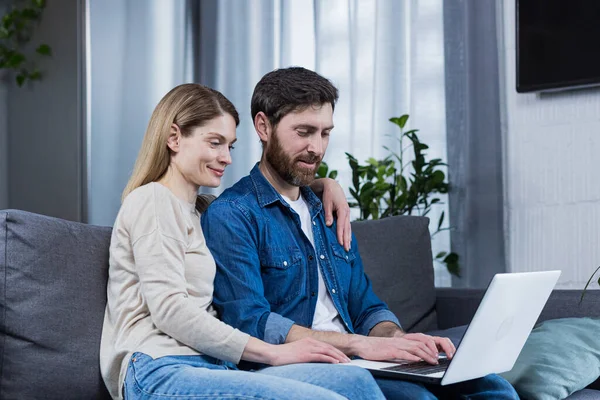 Mutlu genç evli çift evde birlikte oturuyor, kanepede oturuyor gülümsüyor ve dizüstü bilgisayar kullanıyor.