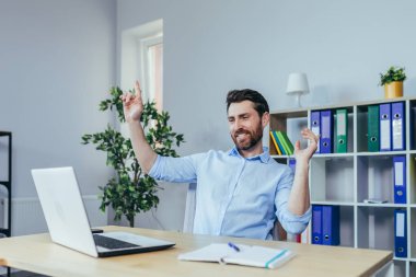 Parlak bir ofiste çalışan bir iş adamı, masada dizüstü bilgisayarla dans etmenin başarısını kutluyor.