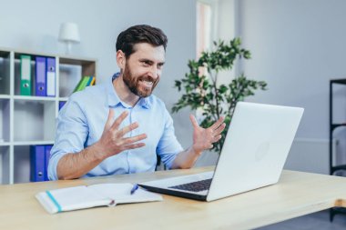 Öfkeli bir işadamı dizüstü bilgisayara bakar ve bağırır, parlak bir ofiste çalışır.