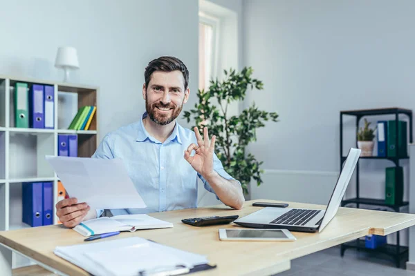 Başarılı bir işadamı kameraya bakar ve gülümser, iş raporlarının uzlaşmasını sağlar, kamera jestini gösterir.