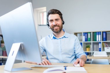 Happy businessman with headset for video call working in modern office, smiling man looking at computer monitor