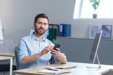 Başarılı sakallı bir işadamı kameraya bakar ve gülümser. Ofisteki bir masada otururken cep telefonu kullanır.
