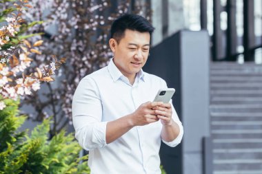 Baharda ofis yakınında Asyalı bir işadamı telefonu kullanır, gömlekli bir serbest çalışan gülümser ve akıllı telefon ekranına bakar.