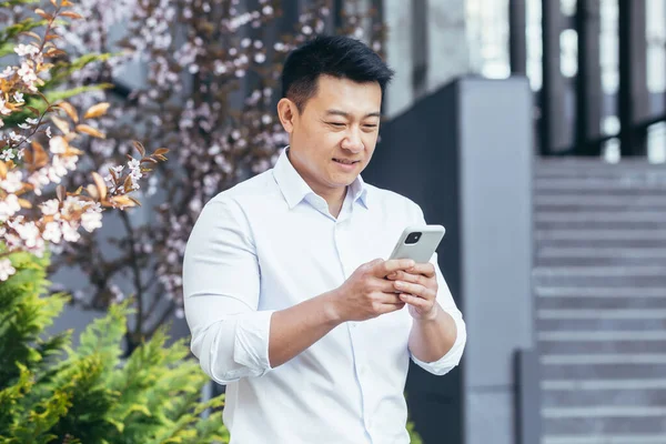Baharda ofis yakınında Asyalı bir işadamı telefonu kullanır, gömlekli bir serbest çalışan gülümser ve akıllı telefon ekranına bakar.