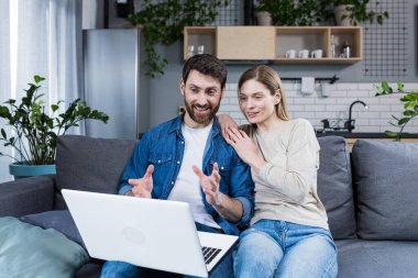 Genç çift, aile, erkek ve kadın evlerinde kanepede oturmuş laptoptan aileye, arkadaşlara video konuşmaları yapıyorlar. El sallarlar, gülümserler, selamlarlar
