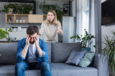 Young family, couple at home, man and woman quarrel, conflict, woman shouting and waving hands. Man sitting on sofa, holding head, listening