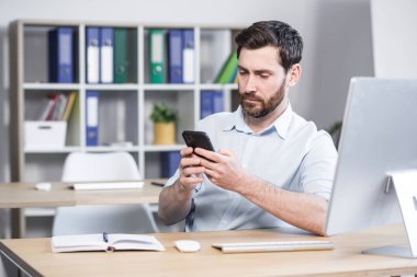 Odaklanmış sakallı genç adam, telefonu kullanıyor, evde masada oturuyor, düşünceli bir şekilde ekrandan okuyor.