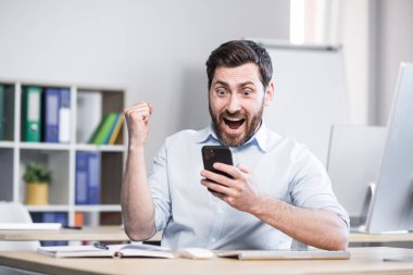 Başarılı serbest çalışan zaferi kutluyor, telefondan iyi haberler okuyor, işadamı ofisinde akıllı telefon kullanıyor.