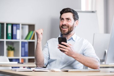 Başarılı serbest çalışan zaferi kutluyor, telefondan iyi haberler okuyor, işadamı ofisinde akıllı telefon kullanıyor.