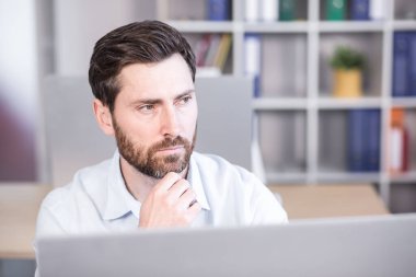 Bir ofis çalışanının portresi. Düşünceli, ciddi, sakallı. Ofisteki bir masada oturan beyaz gömlekli bir bilgisayar..