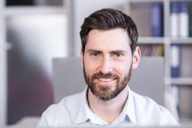 Bir ofis çalışanının portresi. Kameraya bakıyor, gülümsüyor. Ofisteki bir masada oturan beyaz gömlekli bir bilgisayar..