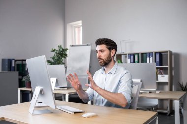 Genç bir adam, bir ofis çalışanı bilgisayarın ofisindeki bir masada oturuyor, bilgisayar kamerasıyla video görüşmesi yapıyor, ortaklarla, müşterilerle iş görüşmesi yapıyor..