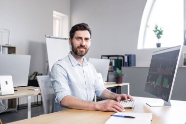 Genç yakışıklı bir adam, ofis çalışanı, grafikle çalışan, masada oturan, klavyede yazan,